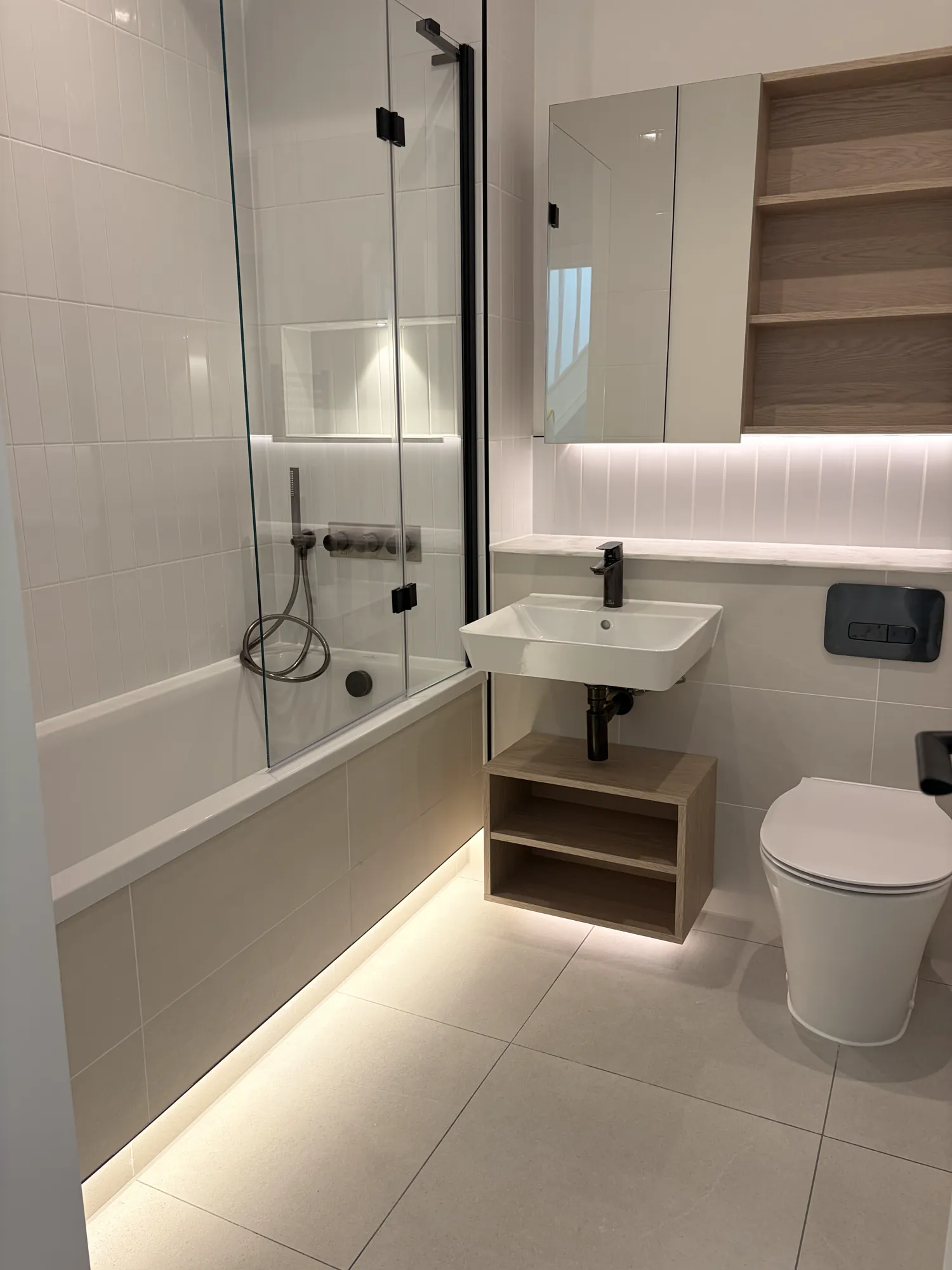Bathroom with wall and floor tiling, a lit shelf recess, an open timber vanity shelf and dark brassware.