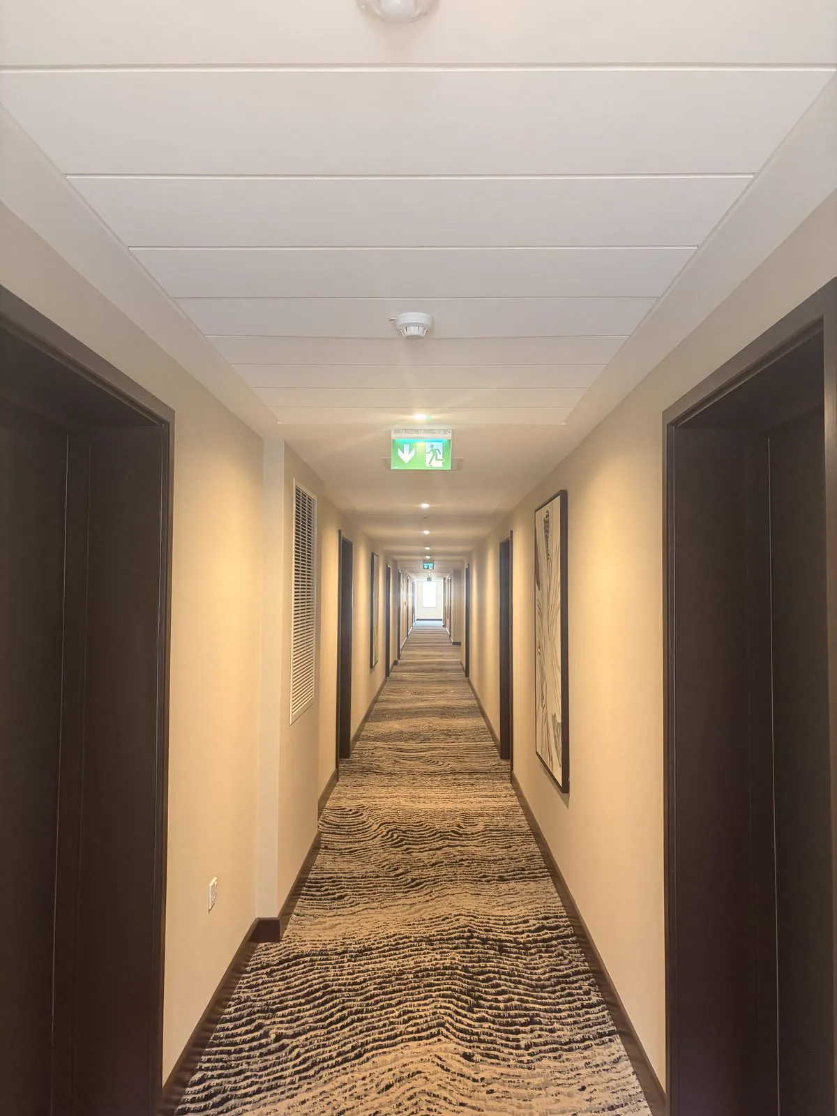 Long apartment-block corridor with cream walls, dark doors, patterned carpet and framed prints, looking towards a window at the far end.