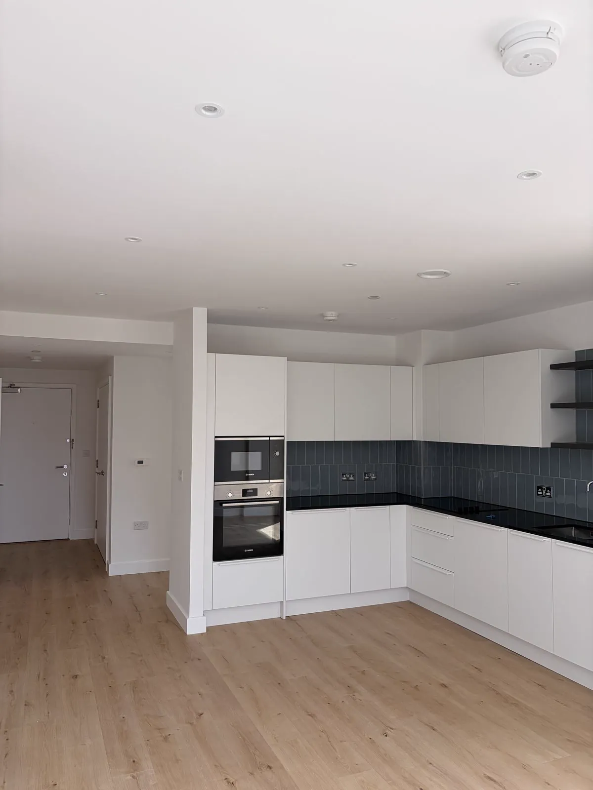 Fitted kitchen with white units, dark worktop, tiled splashback and integrated ovens, with timber-effect flooring and white walls.